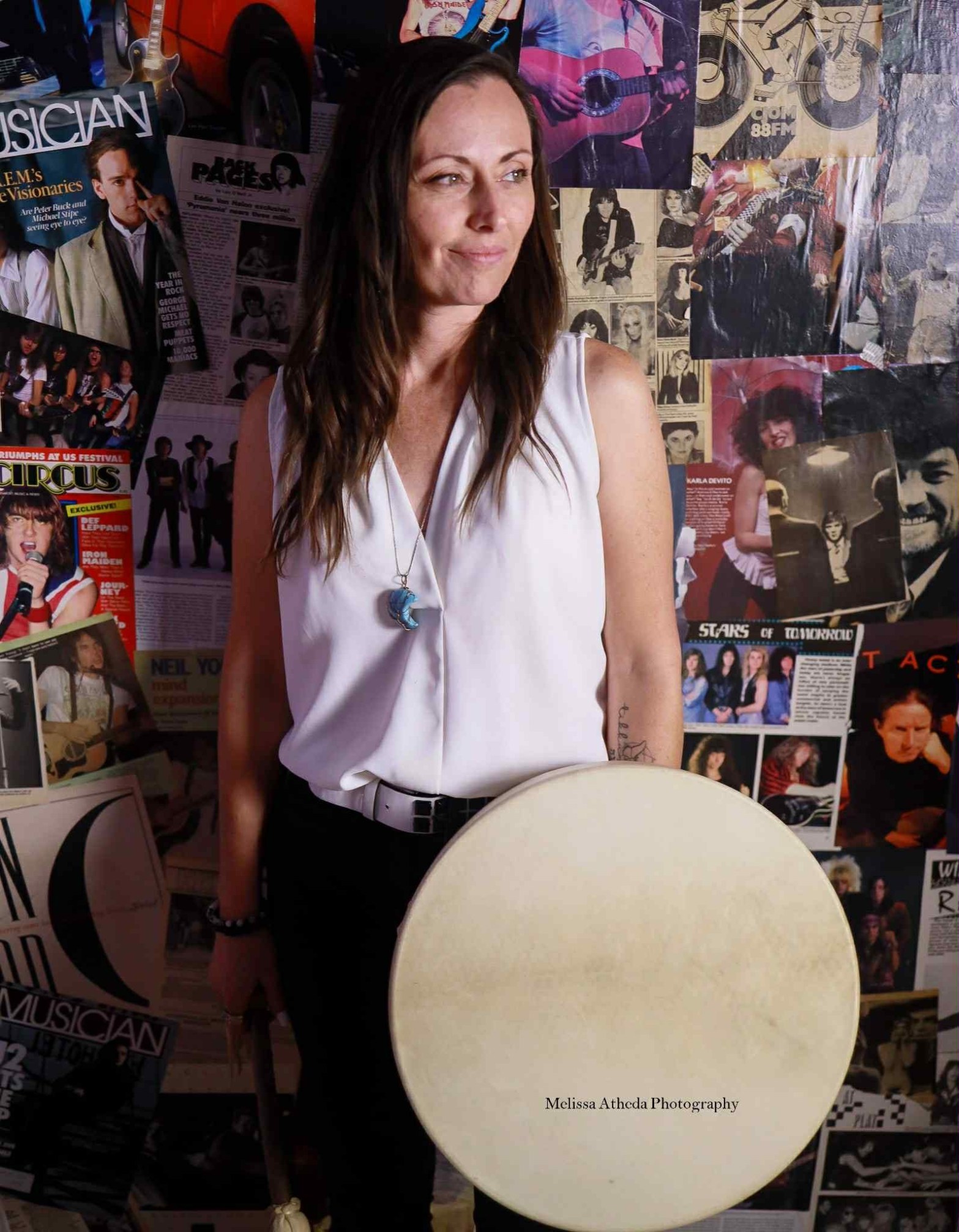 A woman holding a hand drum stands in front of a wall of vintage music posters, looking gently to the side, expressing a grounded presence as she honors ancestral songs and the voices of the women who came before her.
